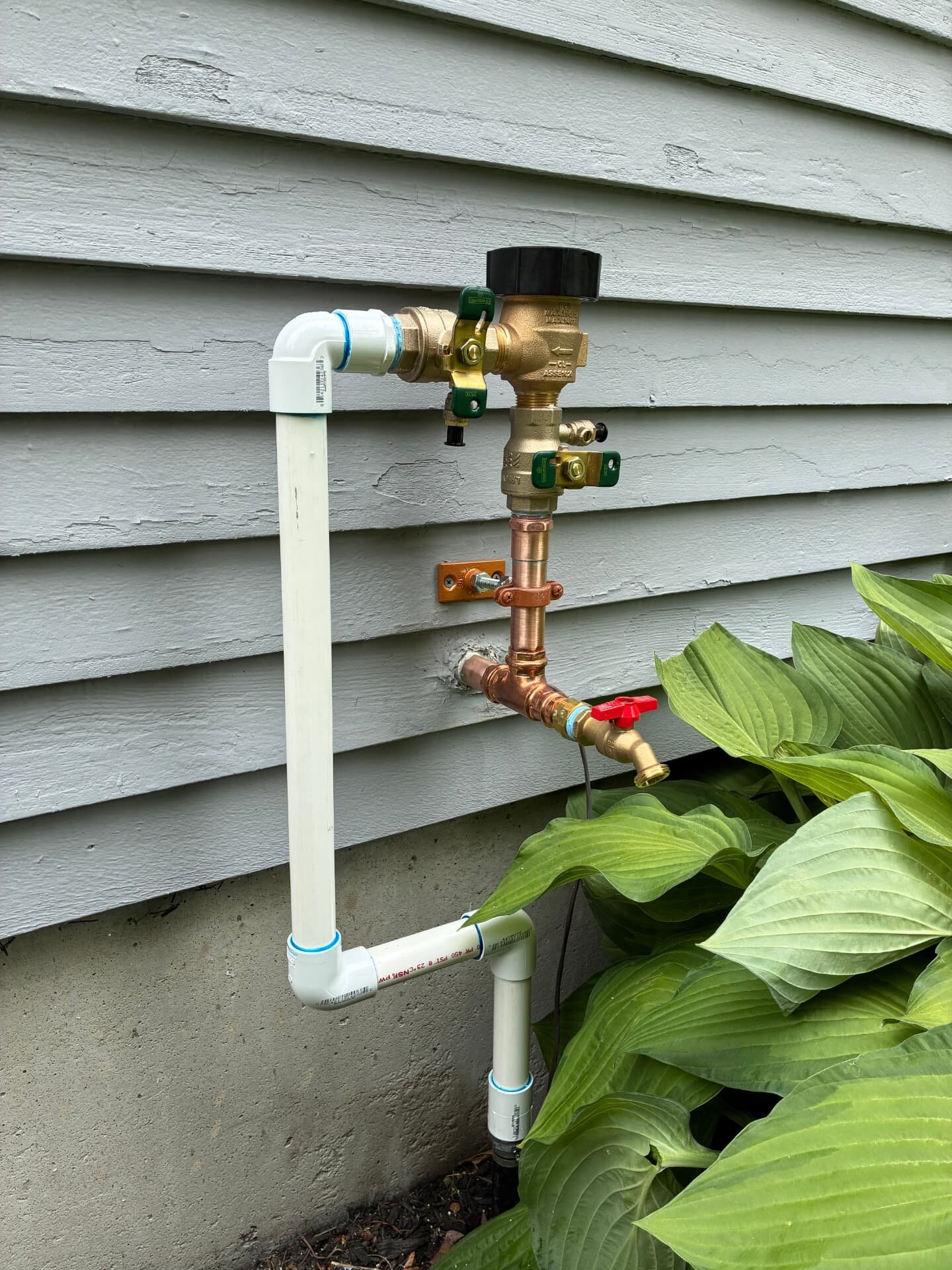 Brass backflow preventer valve with copper and PVC piping installed on a home exterior by Top Shelf Plumbing & Heating